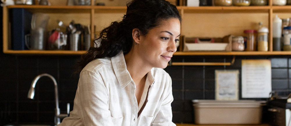 A woman is working on a laptop at the kitchen table, which has a notepad and a pen, a stack of papers, and two slices of cake on a wooden board.