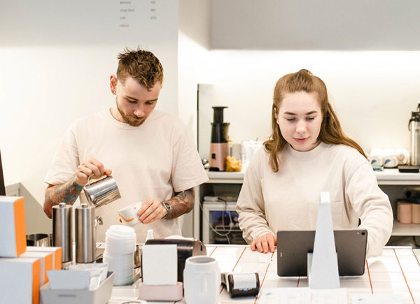 O tânără care privește într-o tabletă și un tânăr care prepară o cafea aflați în spatele tejghelei într-o cafenea.